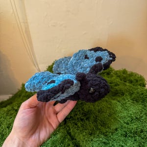May include: A handcrafted crochet butterfly plush toy featuring a dark black body and head with shiny black bead eyes. The wings are constructed from a textured, soft yarn in alternating shades of light blue and dark black, creating a distinct pattern.