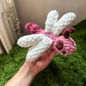 May include: A handmade crochet dragonfly plush toy featuring a soft pink body, dark brown accents on the head and tail, and four textured white wings. The creature has small black bead eyes and a segmented tail with a pink and brown pattern.