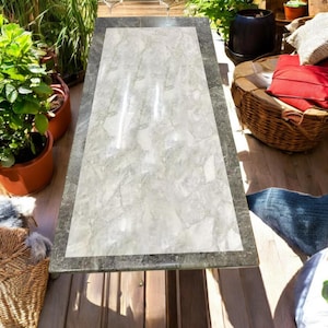 Puede incluir: Una mesa de mármol blanco con un borde gris oscuro. La mesa está hecha de piedra natural y tiene un patrón único. La mesa es perfecta para comer al aire libre o para entretener.