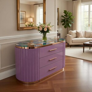 Könnte beinhalten: Eine lila Kommode mit drei Schubladen und einer dekorativen Oberseite. Die Kommode hat eine goldfarbene Umrandung und vertikale Rippendetails an den Seiten. Ein Blumenstrauß steht oben, und ein goldgerahmter Spiegel hängt an der Wand dahinter.