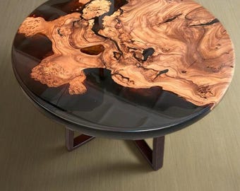 May include: A round wooden table with a black epoxy resin top. The wood has a natural brown color with dark brown grain patterns. The table has a simple black metal base.