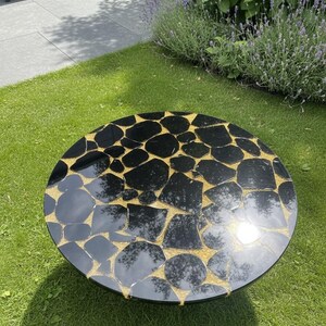 May include: A round outdoor coffee table with a black, mosaic-like top. The mosaic design features irregularly shaped black pieces set against a gold-colored background. The table is placed on a green lawn.