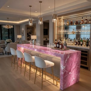 Op de afbeelding: Een luxe thuisbar met een roze marmeren aanrechtblad en gouden accenten. De bar heeft een spiegelachterwand met planken vol drankflessen en glazen. Witte, gestoffeerde barkrukken met gouden poten staan voor de bar.