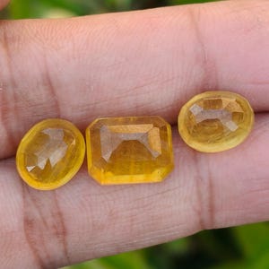 May include: Three faceted yellow gemstones in oval, emerald, and round shapes. The gems are a warm, golden color, with visible internal facets. The gemstones are displayed against a neutral background, highlighting their clarity and brilliance.