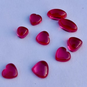 Puede incluir: Colección de gemas brillantes de color magenta, en forma de corazón y ovaladas. Las gemas están dispersas sobre una superficie blanca, con tamaños y formas variadas. Las piedras en forma de corazón son las más prominentes.