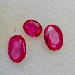 Può includere: Tre gemme ovali sfaccettate in una vibrante tonalità rosa intenso. Le gemme sono esposte su uno sfondo neutro bianco sporco, mostrando la loro chiarezza e brillantezza. Queste pietre lucide sono probabilmente rubini, perfette per la gioielleria.