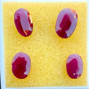 May include: Four oval-cut, faceted rubies presented on a yellow, textured surface. The gemstones are a deep red hue, with internal reflections visible. The rubies are arranged in a square, showcasing their polished surfaces and intricate cuts.