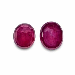 Può includere: Due rubini ovali e sfaccettati. Le pietre preziose sono di un colore rosso intenso e ricco, con una leggera variazione di tonalità tra le due. Le sfaccettature catturano la luce, creando un effetto scintillante. I rubini sono su sfondo bianco.