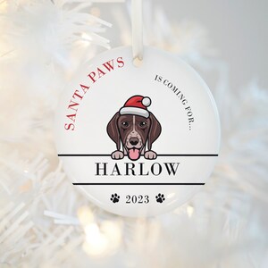 May include: White ceramic Christmas ornament with a cartoon dog wearing a Santa hat. The ornament says "Santa Paws is coming for... Harlow 2023".