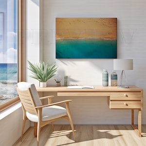 May include: A framed canvas print depicting an aerial view of a beach with turquoise water and golden sand. The artwork hangs above a wooden desk with a lamp, decorative bottles, and a chair, creating a serene office setting.