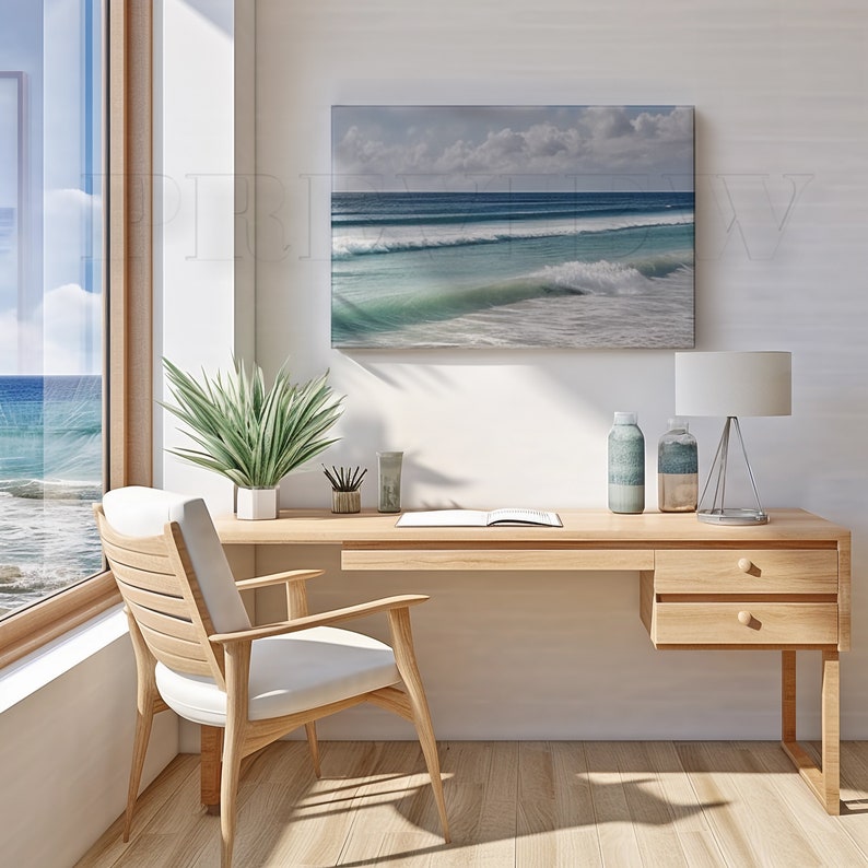 May include: A light wood desk with a white chair, a framed ocean wave print, and a window with a view of the ocean. The desk has a lamp, two decorative bottles, and a plant. The room is bright and airy.