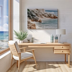 May include: A light wood desk with a matching chair, a framed coastal landscape, and a window with a view of the ocean. The desk has a lamp, decorative bottles, and a plant. The chair has a white cushion.