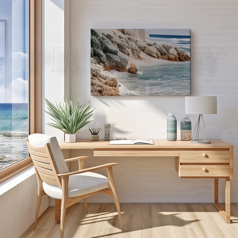 May include: A light wood desk with a matching chair, a framed coastal landscape, and a window with a view of the ocean. The desk has a lamp, decorative bottles, and a plant. The chair has a white cushion.