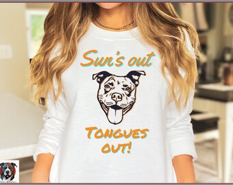 Puede incluir: Sudadera blanca con una ilustración en blanco y negro de un perro con la lengua fuera. El texto "Sun's out Tongues out!" está impreso en naranja y azul en la sudadera.