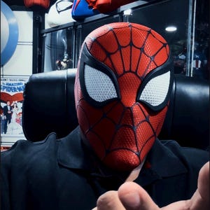 May include: A red and black Spider-Man mask with white eye lenses. The mask has a web pattern and is part of a collection of superhero masks. Other masks are visible in the background.