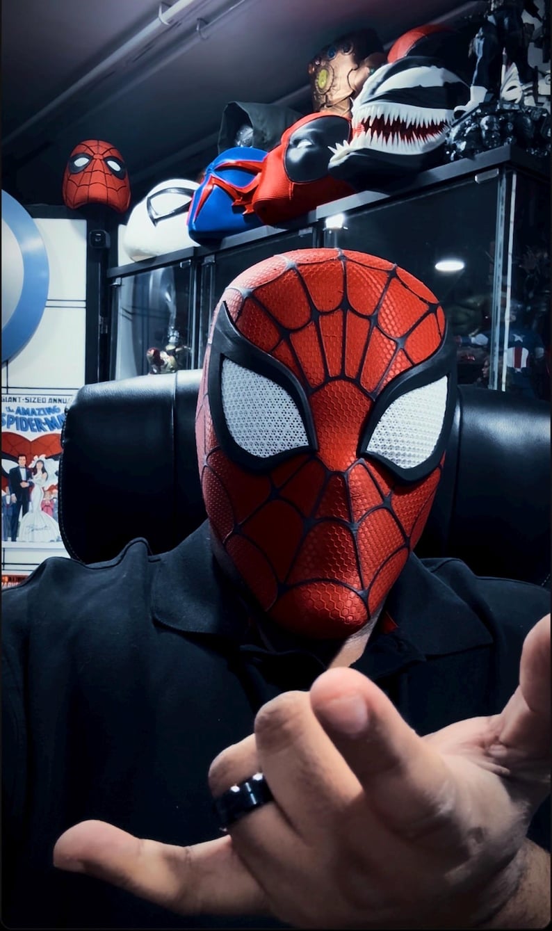 May include: A red and black Spider-Man mask with white eye lenses. The mask has a web pattern and is part of a collection of superhero masks. Other masks are visible in the background.