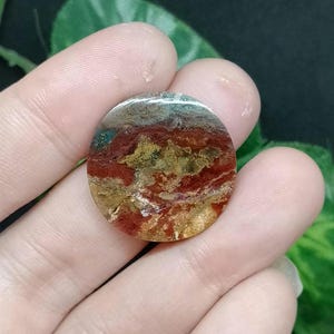 May include: A round, polished agate cabochon with a swirling pattern of red, brown, gold, and green colors.