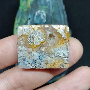 Puede incluir: Un cabujón de ágata pulido de forma cuadrada con un patrón blanco, amarillo y gris. La piedra tiene un aspecto natural y orgánico.