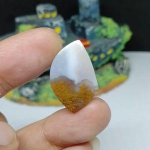 Puede incluir: Un cabujón de ágata blanco y marrón con forma de lágrima. La ágata tiene una parte superior blanca con patrones marrones y blancos en la parte inferior.