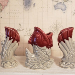 May include: Three ceramic vases in the shape of fish. The vases are decorated with a red and white glaze. The fish are in different poses, with one swimming, one standing upright, and one with its tail curled up.