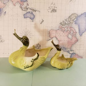May include: Two green and brown glazed ceramic swan planter pots. The larger swan has its head turned to the left and the smaller swan has its head turned to the right. Both swans have a cream colored interior.