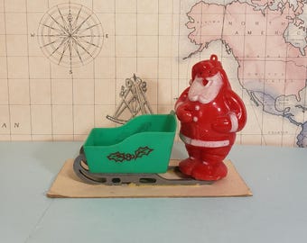 May include: A red Santa Claus figurine with a white beard and trim stands next to a green sleigh decorated with holly. The Christmas-themed decorations are on a cardboard base, set against a vintage map.