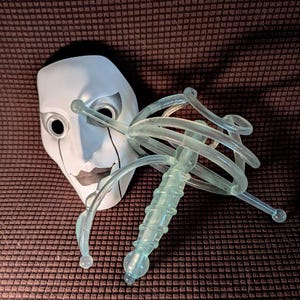 以下が含まれることがあります： 目と唇に黒いディテールが施された白い演劇用マスクと、中央の隆起したロッドの周りに螺旋状のガラスコイルが巻かれ、先端が小さな丸い球体で終わる半透明の淡い緑色のガラス彫刻が並んでいます。