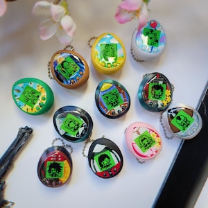 Könnte beinhalten: Eine Sammlung farbenfroher, eiförmiger Digital-Haustierspielzeuge mit Schlüsselanhängern. Jedes Spielzeug hat einen kleinen Bildschirm mit pixeligen Charakteren. Die Spielzeuge sind in verschiedenen Farben erhältlich, darunter Gelb, Grün, Rosa und Schwarz.