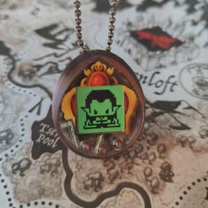 Pode incluir: Um colar com pingente com um personagem de duende verde pixelado dentro de uma resina transparente em forma oval. O duende está emoldurado por uma moldura ornamentada amarela e vermelha, suspensa por uma corrente de bolinhas prateada. O fundo é um padrão semelhante a um mapa.