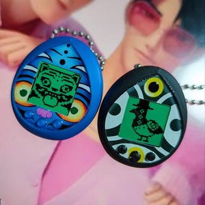 Op de afbeelding: Twee hangerkettingen met retro handheld game ontwerpen. Eén is blauw met een tijgerafbeelding, de andere is zwart met een eendafbeelding. Beide hebben een groen scherm en een zilveren ketting.