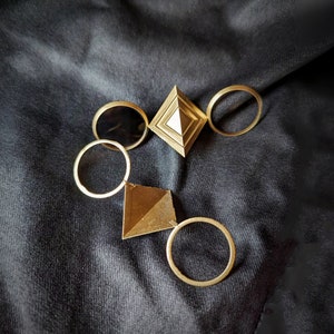 Puede incluir: Una colección de joyas doradas, que incluye anillos y colgantes de formas geométricas, dispuestas sobre una superficie oscura y texturizada. Las joyas incluyen un colgante en forma de diamante y un colgante en forma de pirámide, junto con varios anillos circulares simples.