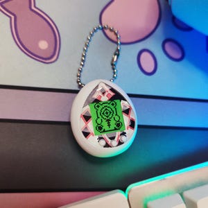 Puede incluir: Un juguete digital para mascotas en forma de huevo blanco con una cadena plateada. La pantalla muestra un gráfico verde de una criatura. El juguete tiene tres botones pequeños debajo de la pantalla y un borde negro, rosa y blanco.