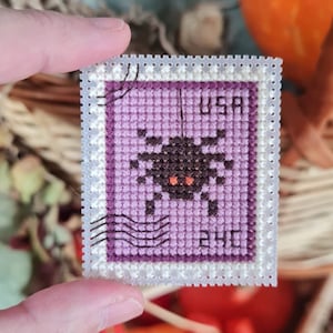 May include: A cross-stitched stamp with a Halloween theme. The stamp features a black spider with orange eyes on a purple background. The stamp reads "USA" and "24p". The stamp is held in a hand.
