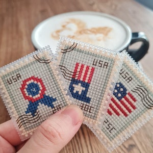 May include: Three rectangular cross-stitch pieces designed to look like vintage postage stamps. Each stamp features red, white, and blue patriotic motifs, including a circular emblem, a flag-inspired design, and a star-spangled pattern with USA text.