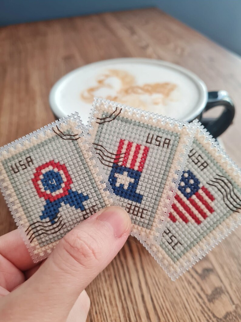 May include: Three rectangular cross-stitch pieces designed to look like vintage postage stamps. Each stamp features red, white, and blue patriotic motifs, including a circular emblem, a flag-inspired design, and a star-spangled pattern with USA text.