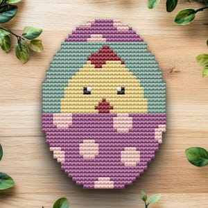 May include: A cross-stitch Easter egg design featuring a yellow chick hatching from a purple egg. The egg has a light green background and a purple top with white polka dots. The chick has black eyes and a red beak.