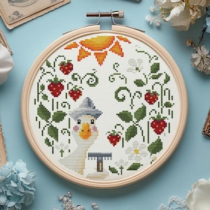 May include: A cross-stitch embroidery project in a round wooden hoop featuring a white fabric background. The design depicts a central goose with a blue hat, surrounded by red strawberries, green leaves, white flowers, and an orange sun.
