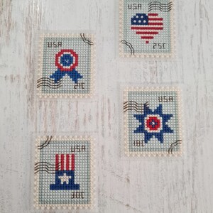 May include: Four rectangular cross-stitch pieces designed to resemble postage stamps. Each features a light blue background, the text USA, and black cancellation marks. Designs include a red, white, and blue ribbon, a heart, a star, and a top hat.
