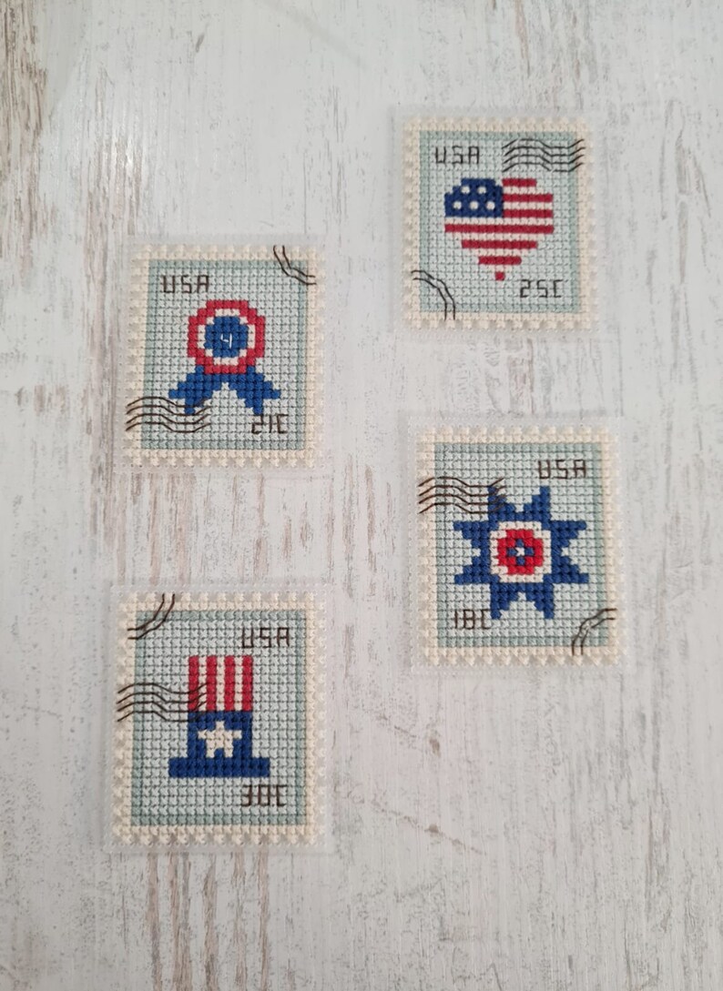 May include: Four rectangular cross-stitch pieces designed to resemble postage stamps. Each features a light blue background, the text USA, and black cancellation marks. Designs include a red, white, and blue ribbon, a heart, a star, and a top hat.
