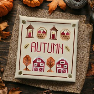 May include: A square cross-stitch sampler on cream fabric featuring the word AUTUMN in red letters. The design includes two lanterns, two baskets, two orange trees, and two red barns with white roofs, all bordered by a thin brown stitched frame.