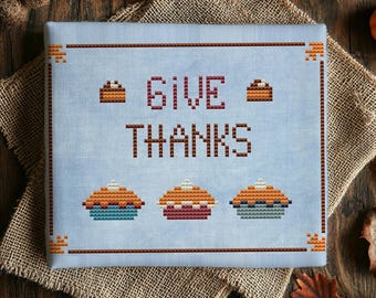 May include: A light blue cross-stitch fabric piece featuring the text GIVE THANKS in dark red block letters. The design includes two small pie icons above the text and three larger pie icons below, all framed by a dark red border.