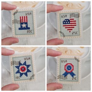 May include: A set of four rectangular cross-stitch pieces designed to resemble postage stamps. Each piece features a light blue background, the text USA, black postmark lines, and patriotic motifs including a top hat, heart, star, and ribbon.