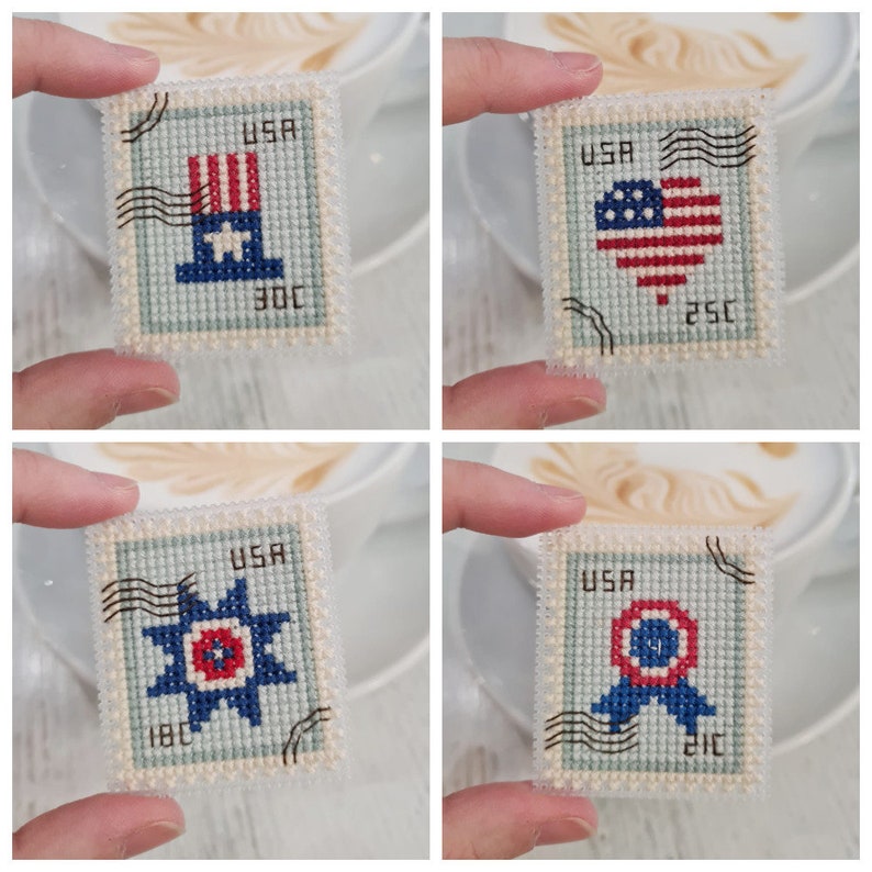 May include: A set of four rectangular cross-stitch pieces designed to resemble postage stamps. Each piece features a light blue background, the text USA, black postmark lines, and patriotic motifs including a top hat, heart, star, and ribbon.