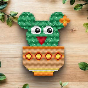 May include: A cross-stitched cactus design in a terracotta pot. The cactus is green with a smiling face, and the pot has a decorative pattern. The design is set against a wooden background, creating a playful, handcrafted aesthetic.
