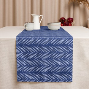 Può includere: Un runner da tavolo blu con un motivo a chevron bianco. Il runner è su una tovaglia beige e ci sono due tazze in ceramica bianca e un'anfora bianca sul tavolo.