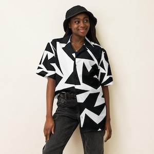 Puede incluir: Una camisa de manga corta con un estampado geométrico en blanco y negro. El diseño presenta formas negras audaces sobre un fondo blanco. La camisa se combina con vaqueros negros y un sombrero de pescador negro, creando un fuerte contraste visual.