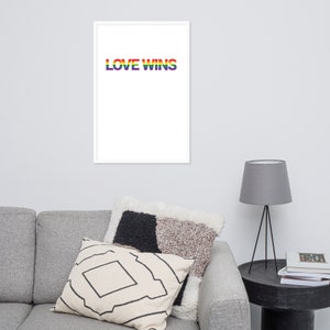 Puede incluir: Un cuadro enmarcado con las palabras "LOVE WINS" en colores del arcoíris sobre un fondo blanco. El cuadro se exhibe en una pared sobre un sofá gris con cojines decorativos y una mesa auxiliar con una lámpara.