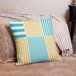 May include: Square cushion with a patchwork design. It features sections of turquoise stripes, white stripes, and yellow zigzags. The cushion is on a bed with a striped blanket and a woven cushion.