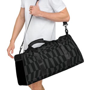 Könnte beinhalten: Schwarz-graue Reisetasche mit geometrischem Muster. Die Tasche hat einen Schultergurt und einen Griff. Die Tasche besteht aus strapazierfähigem Material und hat einen Reißverschluss. Die Tasche ist ca. 51 cm lang, 25 cm breit und 25 cm hoch.