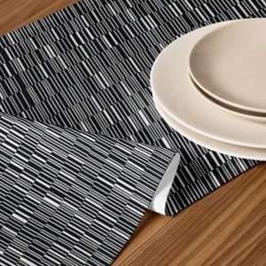 May include: Black and white geometric patterned table runner with a white border. The runner is folded in half and placed on a wooden table with two white plates stacked on top of each other.